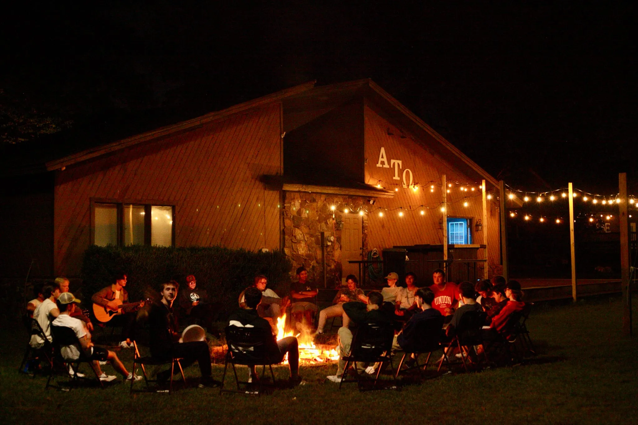 The Beta Tau chapter gathered around a bonfire at night in front of the lodge, ATO lettering lit on the gable