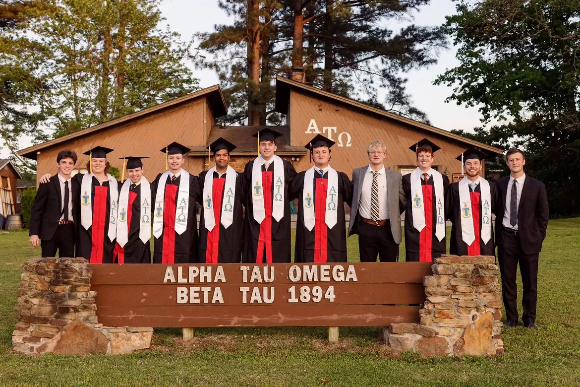 Graduating seniors in caps and gowns with ATO stoles in front of the lodge sign