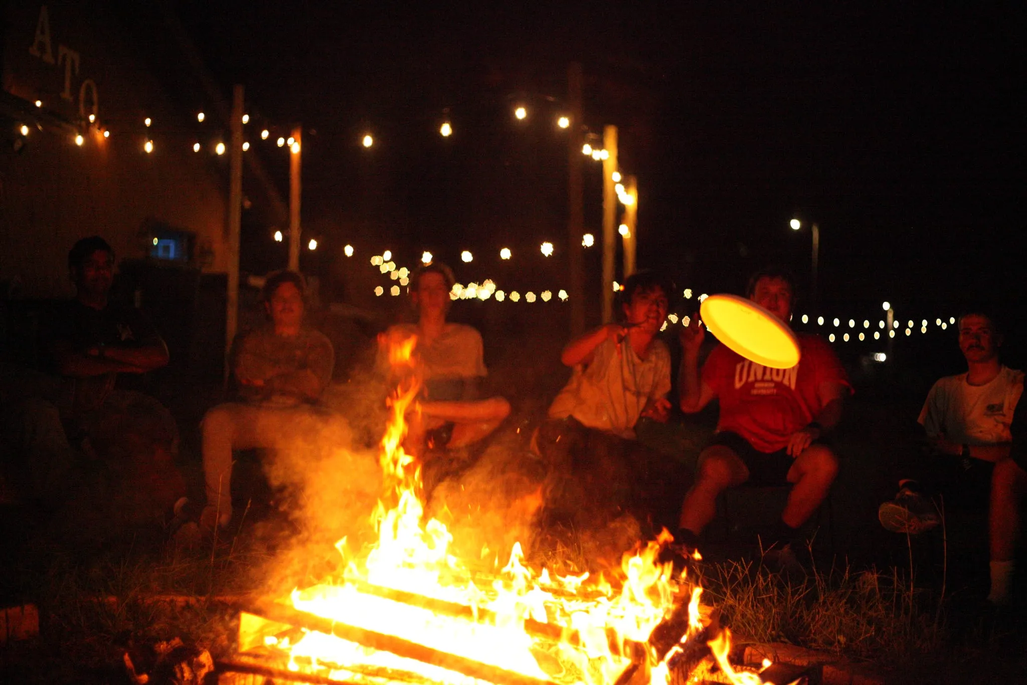Brothers gathered around a bonfire at night with string lights overhead